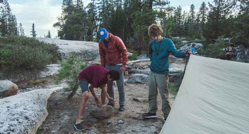 Three students work to set up a tarp shelter in a wooded area. 